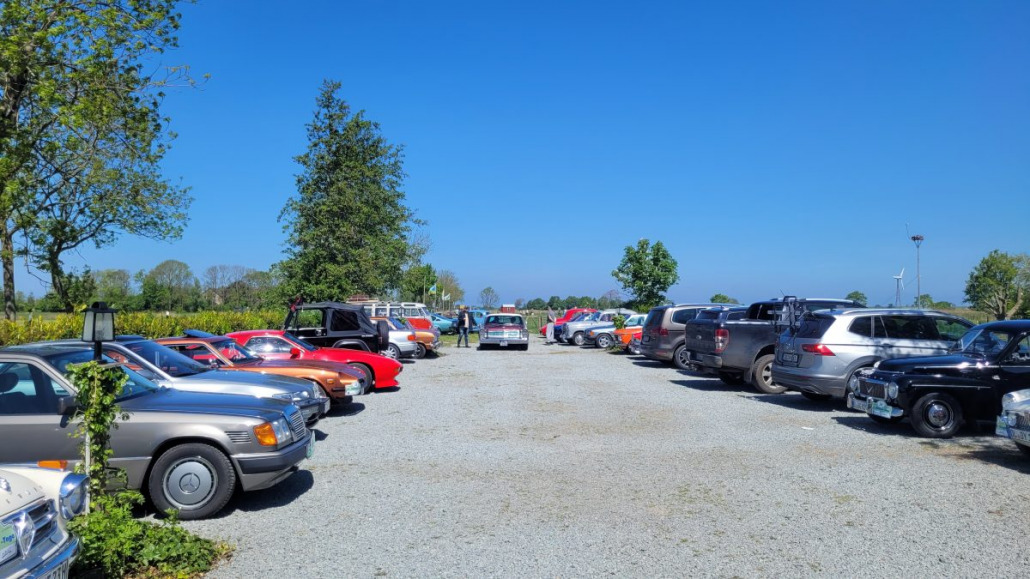 Ein geschotterter Parkplatz voller verschiedener Autos, darunter auch Oldtimer, unter einem klaren blauen Himmel mit Bäumen und Feldern im Hintergrund.